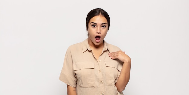 Pretty young woman looking shocked and surprised with mouth wide open, pointing to self