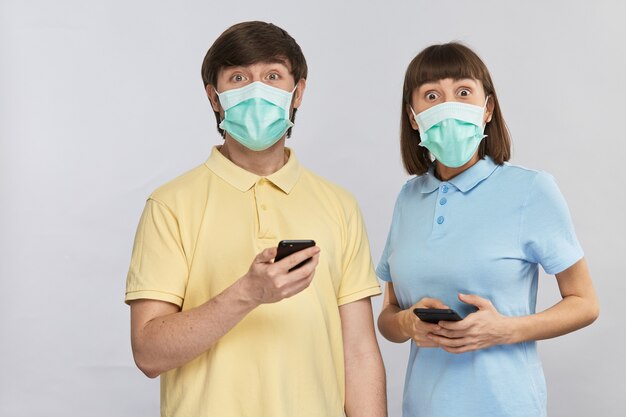 Pretty young woman and man in yellow and blue shirts holding smartphones and surprisely looking to camera with big eyes in protection masks, copy space