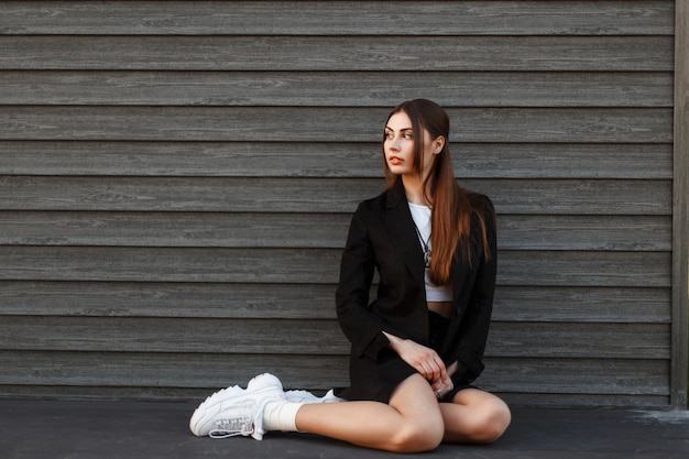 Pretty young woman model in a fashion black coat with white sneakers sitting near a wooden wall