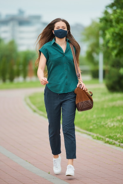 A pretty young woman in a navy blue medical face mask jumps holding a leather bag in the park