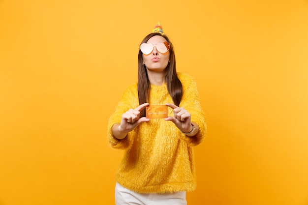 Pretty young woman in orange heart glasses birthday party hat blowing lips holding credit card, enjoying holiday, celebrating isolated on yellow background. People sincere emotions, lifestyle concept.