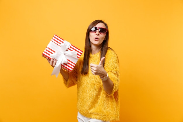 Pretty young woman in red eyeglasses blowing lips showing thumb up holding red box with gift present isolated on bright yellow background. People sincere emotions, lifestyle concept. Advertising area.