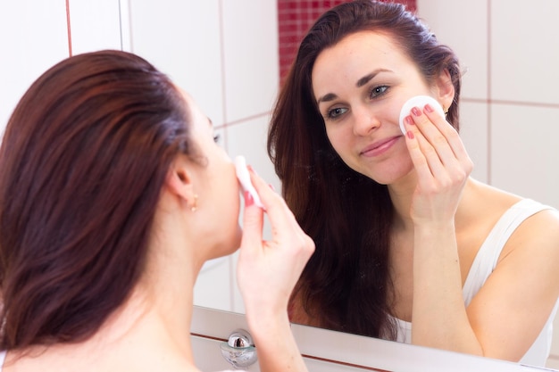 Pretty young woman in shirt using face cream in front of the mirror in her bathroom