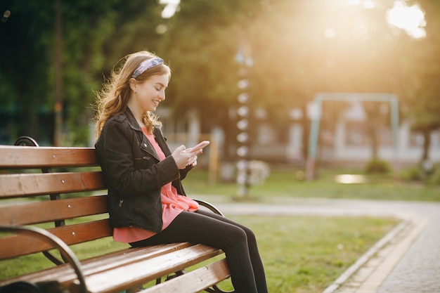 Pretty young woman sits on a bench in the city park, actively texts messages via her phone