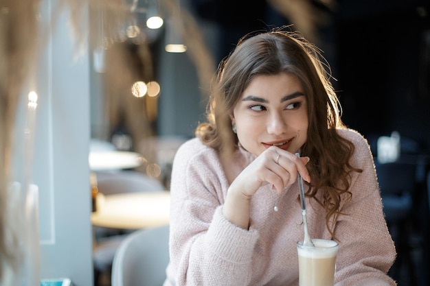 사진 카푸치노 한 잔과 긴 숟가락을 손에 들고 카페에 앉아 있는 예쁜 젊은 여성