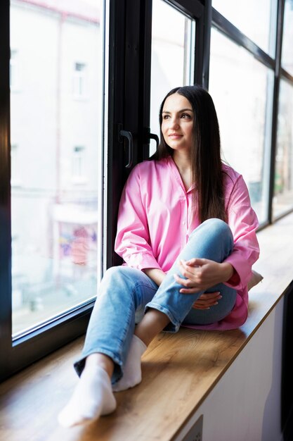 Pretty young woman sitting on windowsill relaxing at home looking at camera Smiling calm lady chilling in apartment dreaming thinking of peaceful time enjoying peace of mind