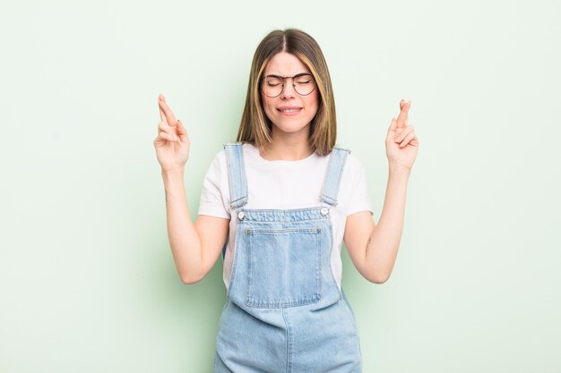 Pretty young woman smiling and anxiously crossing both fingers feeling worried and wishing or hoping for good luck