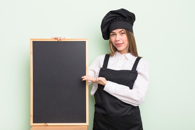 Pretty young woman smiling cheerfully feeling happy and showing a concept chef with blackboard