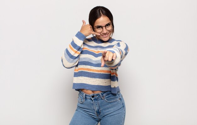 Pretty young woman smiling cheerfully and pointing to camera while making a call you later gesture, talking on phone