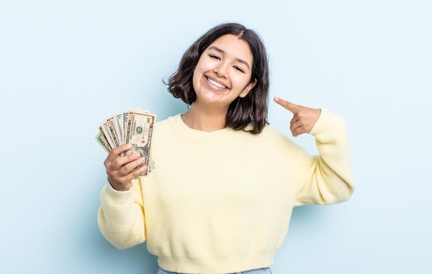 Pretty young woman smiling confidently pointing to own broad smile. dollar banknotes concept