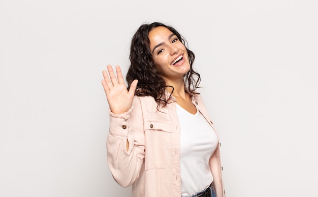 Pretty young woman smiling happily and cheerfully, waving hand, welcoming and greeting you, or saying goodbye