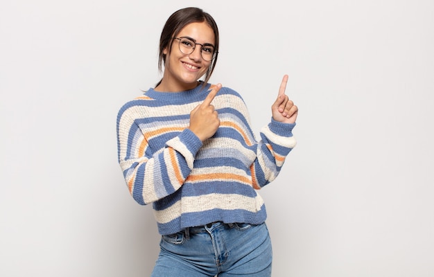 Pretty young woman smiling happily and pointing to side and upwards with both hands showing object in copy space