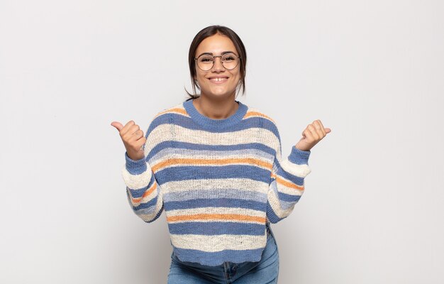 Pretty young woman smiling joyfully and looking happy, feeling carefree and positive with both thumbs up