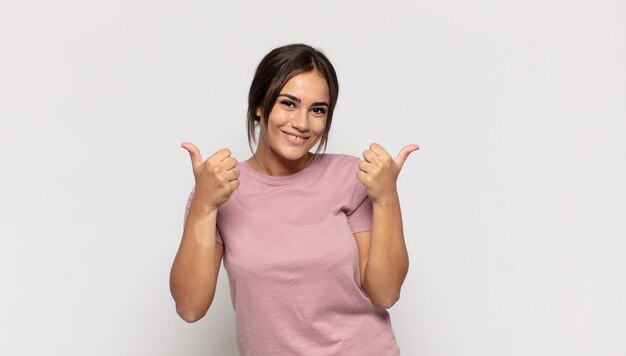 Pretty young woman smiling joyfully and looking happy, feeling carefree and positive with both thumbs up