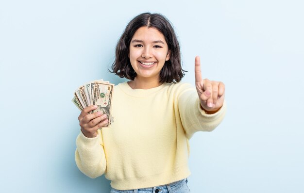 Pretty young woman smiling and looking friendly, showing number one. dollar banknotes concept