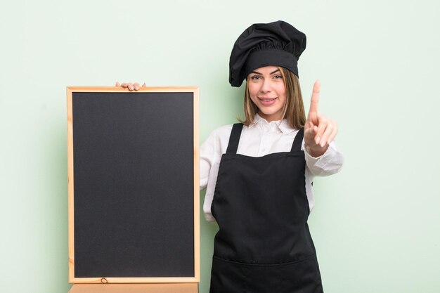 Pretty young woman smiling proudly and confidently making number one chef with blackboard