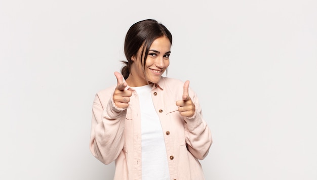 Pretty young woman smiling with a positive, successful, happy attitude pointing to the camera, making gun sign with hands