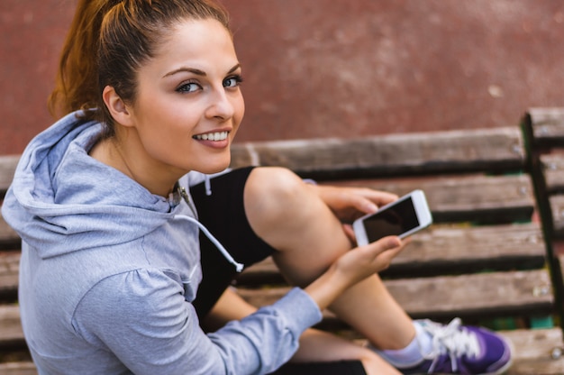 Pretty young woman in sporty outfit holding a smartphone in the hands while sitting on a park bench.