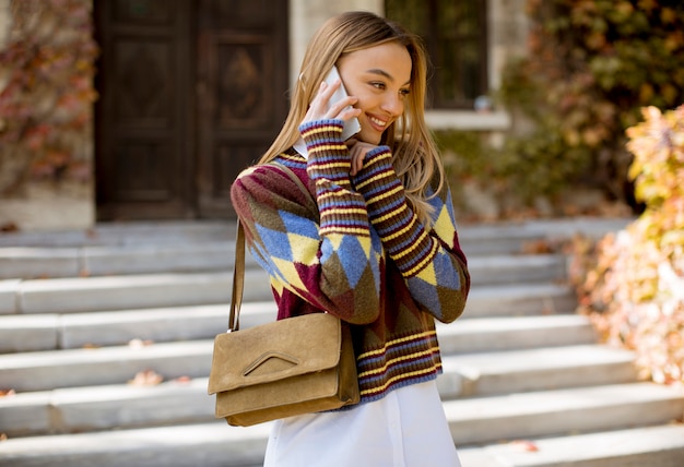 Pretty young woman standing with mobile phone on street at autumn day