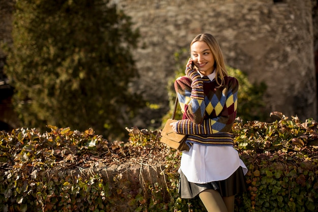 Pretty young woman standing with mobile phone on street at sunny autumn day