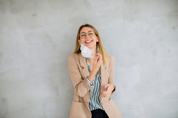 Pretty young woman standning by the grey wall and taking off a\
respiratory mask from coronavirus disease