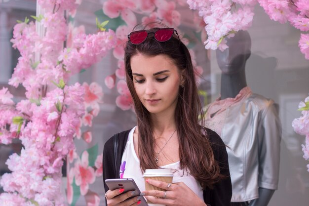 Pretty young woman stands near a storefront keeps the coffee and stares at the phone