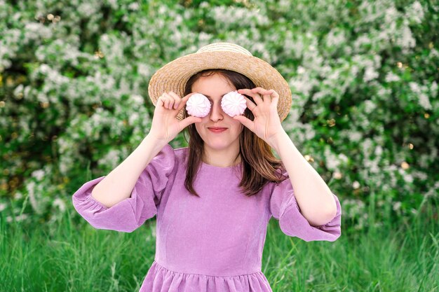 A pretty young woman in a straw hat poses with marshmallows at a picnic in a blooming apple orchard