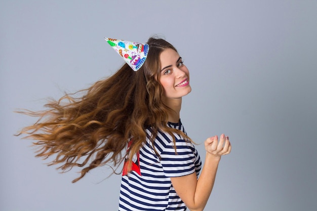 Bella giovane donna in maglietta spogliata e berretto celebrativo che gira su sfondo grigio in studio