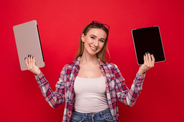 A pretty young woman in stylish clothes holds a tablet and a laptop on a red background