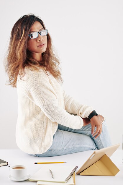 Pretty young woman in sweater sitting on desk with textbooks and digital tablet after having online lesson