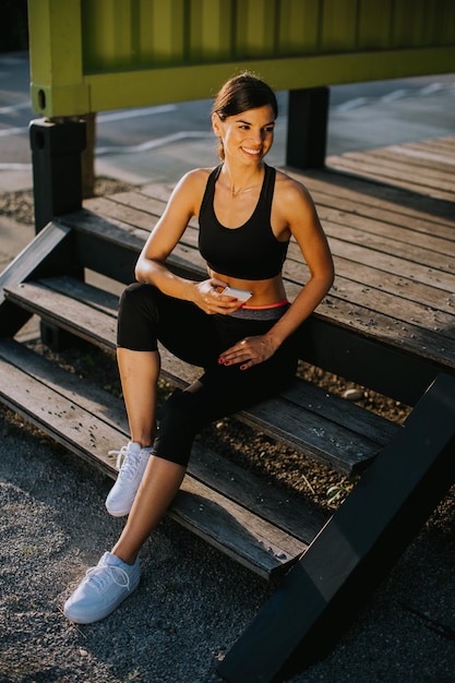 Pretty young woman taking a break during exercising outside and\
using mobile phone