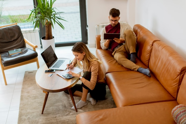 Photo pretty young woman using laptop and young man using digital tablet while sitting on sofa at home