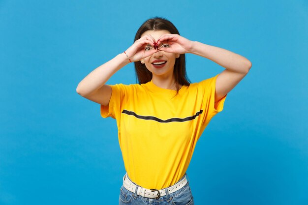 Pretty young woman in vivid casual clothes looking camera holding hands near eyes imitating glasses or binoculars isolated on bright blue wall background. People lifestyle concept. Mock up copy space.