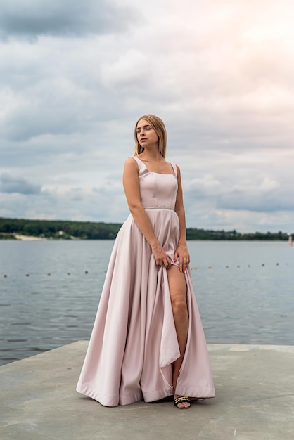 공원의 호수 근처에서 포즈를 취하는 분홍색 패션 이브닝 드레스를 입은 예쁜 젊은 여성