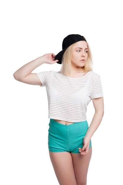 Довольно молодая женщина в зеленых шортах, полосатой футболке и кепке позирует изолированно на белом фоне