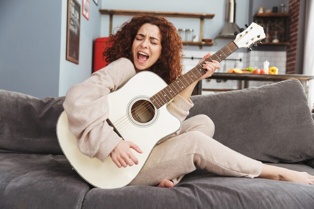 pretty young woman wearing house clothes playing acoustic guitar while sitting on sofa in apartment