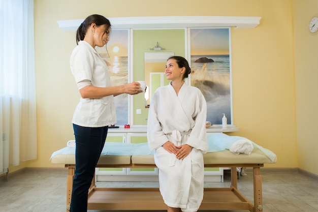 Pretty young woman wearing robe and masseuse talking after massage in a spa drinking some tea