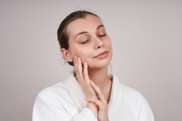 A pretty young woman in a white robe touches the delicate skin of her cheek, closing her eyes. a light wall. Facial care concept.