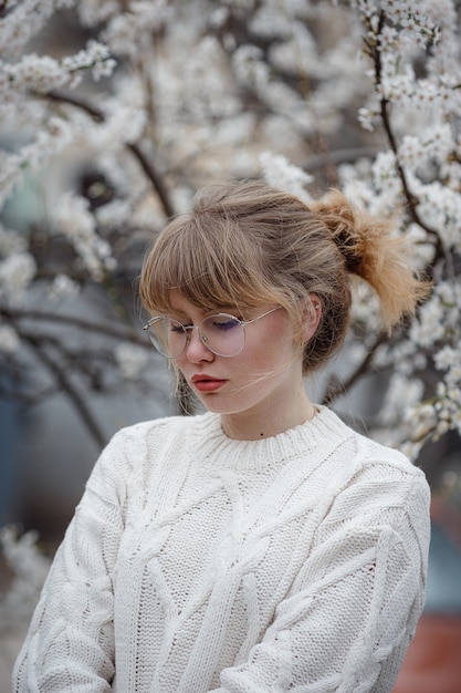 Piuttosto giovane donna in un maglione bianco nel giardino di fiori di ciliegio, tempo primaverile. foto di moda all'aperto