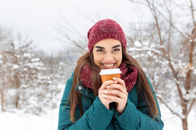 Bella giovane donna in abito invernale con in mano una tazza usa e getta piena di caffè o tè caldo ragazza con in mano una tazza di bevanda calda e passeggiate all'aperto in inverno