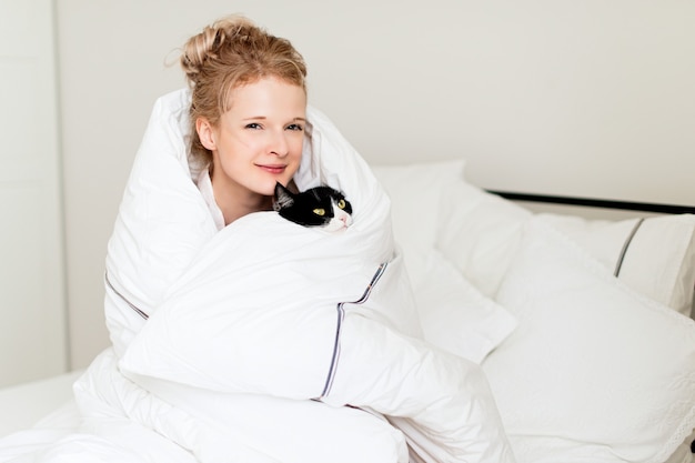 Pretty young woman with black cat in blanket in white bedroom