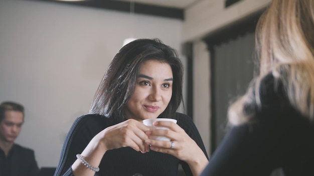 Pretty young woman with black hair drinking coffee and talking with girlfriend in the cafe, portrait