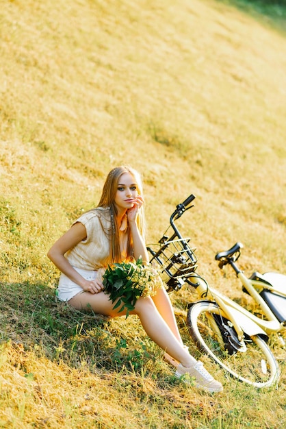 野生の花の花束を持つかなり若い女性が夏休みを自転車に乗るために草の上に座っています