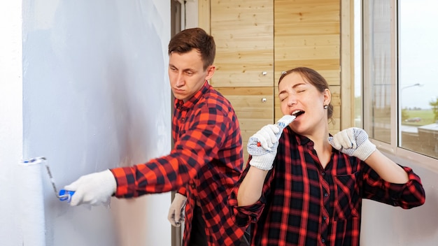 Pretty young woman with brush sings and dances while tired man paints wall with roller repairing light spacious glazed loggia at home