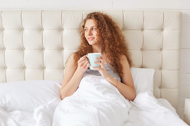 Pretty young woman with curly hair has morning aromatic coffee in bed