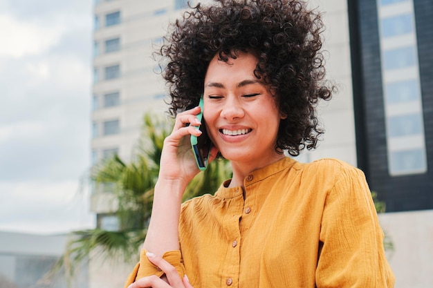 巻き毛と黄色いシャツを着たかなり若い女性が、スマートフォンを使って電話をかけ、幸せそうな顔をして携帯電話で話すヒスパニック系の笑顔の女の子通信のコンセプト