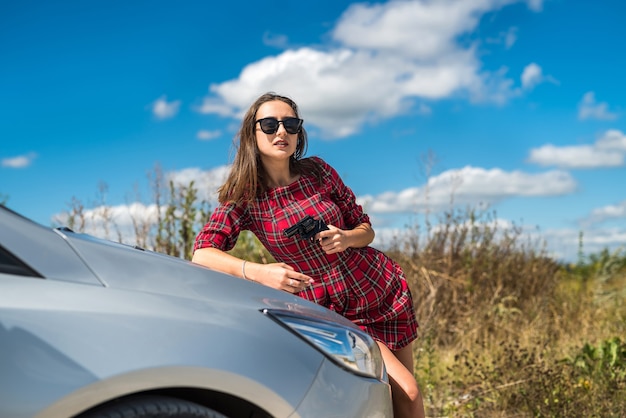 Pretty young woman with gun near her car at nature, crime concept