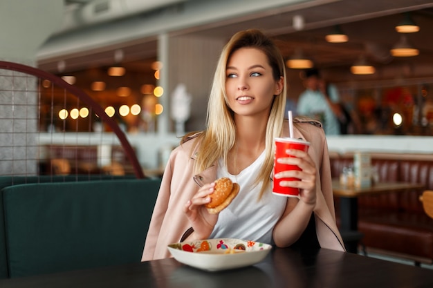 ハンバーガー、フライドポテトと屋内でファーストフードを食べる飲み物を持つかなり若い女性