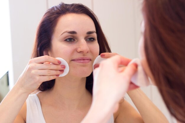 Foto bella giovane donna con lunghi capelli scuri in camicia bianca che rimuove il trucco con tamponi di cotone davanti allo specchio nel suo bagno