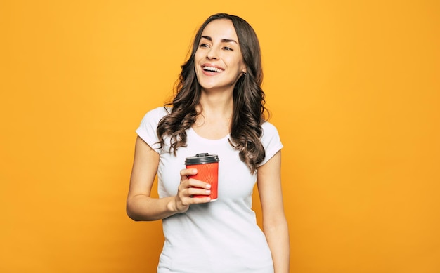 Pretty young woman with long wavy dark hair and eye catching smile on her face is holding a cup of hot tasty coffee in her hands